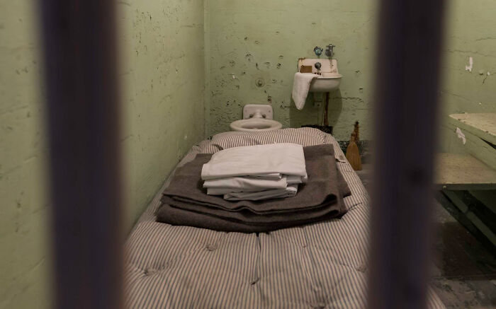 View of a sparse psychiatric facility room with bed, sink, and striped mattress behind bars
