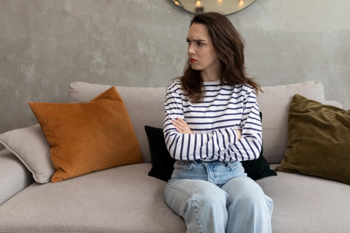 A woman with a furrowed brow and arms crossed sits on a couch, looking disgruntled, learning to defend herself.