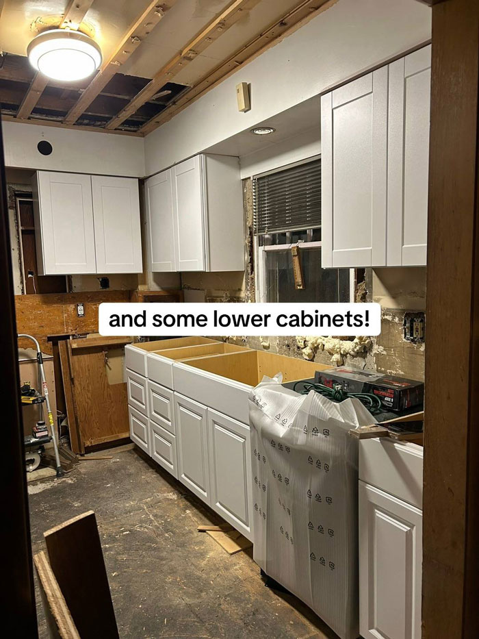 A kitchen mid-renovation with white upper and lower cabinets installed, walls exposed, and a messy floor, showing a contractor's unfinished work.