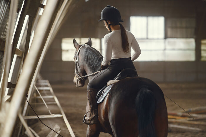 Young woman on a horse, viewed from behind in an arena. A person crossing to the other side with her horse.