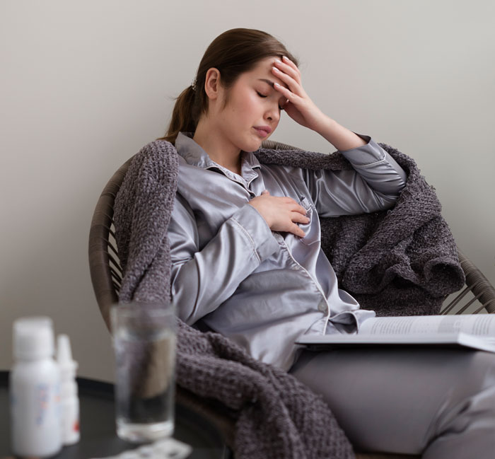 Woman in pajamas, wrapped in a blanket, looking ill with hand on forehead. A side table holds medication and water. Wrong facts can make you sick.