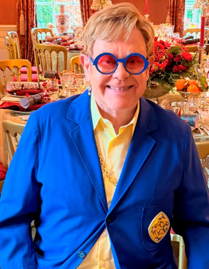 Elton John smiling, wearing a blue blazer and unique glasses, demonstrating how celebrities can be frugal.