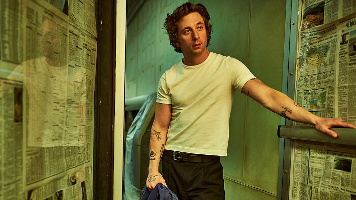 Jeremy Allen White como Carmen Berzatto em The Bear, encostado em uma parede coberta de jornal, parecendo pensativo. O Urso Temporada 5.