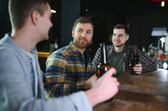 Three men at a bar, sharing drinks and smiling, perhaps recalling cringe situations causing second-hand embarrassment.