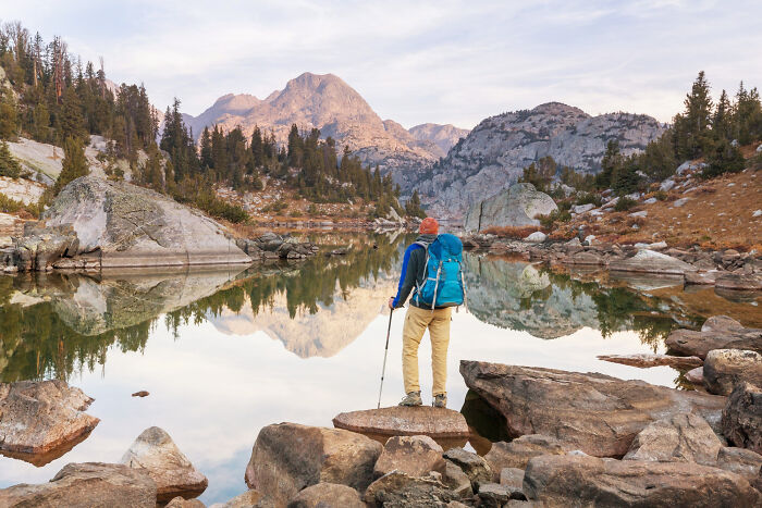 A lone tourist with a blue backpack and hiking poles stands on rocks overlooking a serene mountain lake.