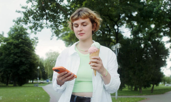Person holding ice cream cone and smartphone standing in park