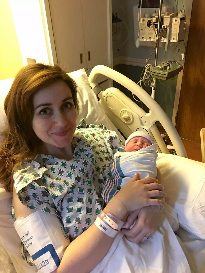 A new mother smiles in a hospital bed, holding her swaddled newborn. This image highlights doctors' awesome sense of humor.