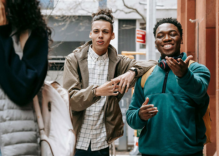 Three young men outdoors. Two are interacting, one smiling with headphones, perhaps displaying actions that people of low intelligence do.