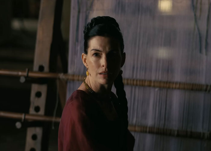 Anne Hathaway in a red dress with a braided hairstyle. Her face is partially shadowed, capturing her acting scene for The Odyssey.