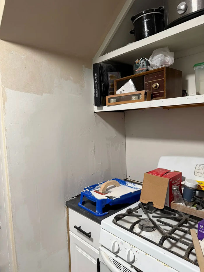 Kitchen wall with visible contractor imperfections, showing a paint tray and roller on the counter, next to a stove.