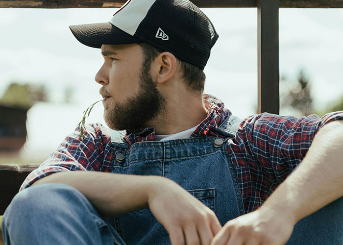 Young man in a baseball cap, plaid shirt, and overalls with a straw in his mouth, dispelling US stereotypes.