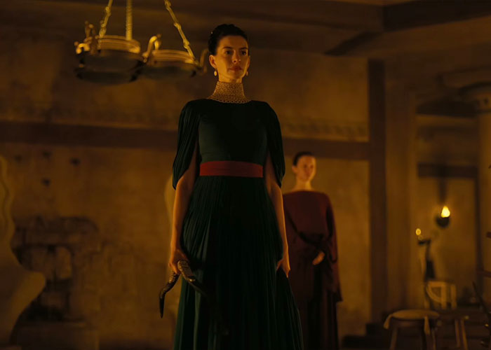 Anne Hathaway in The Odyssey, wearing a green dress with a red belt, holding a dagger, showcasing her acting.