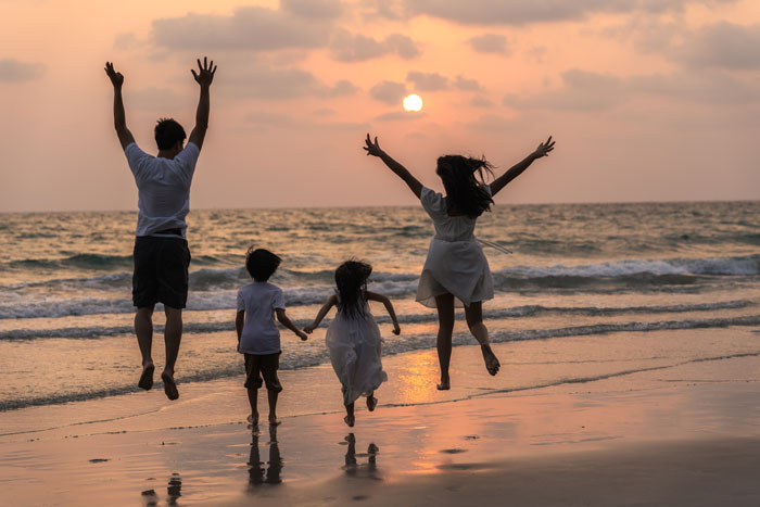 A family of four, including two children, jumping for joy on a beach at sunset, embodying simple luxury.