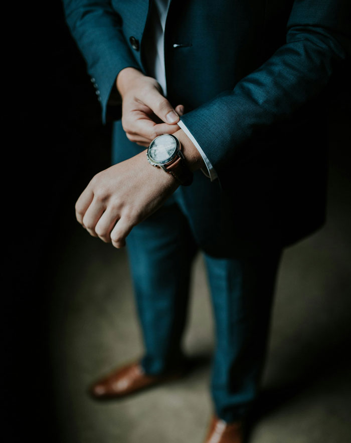 Man adjusting watch on wrist wearing blue suit and brown shoes