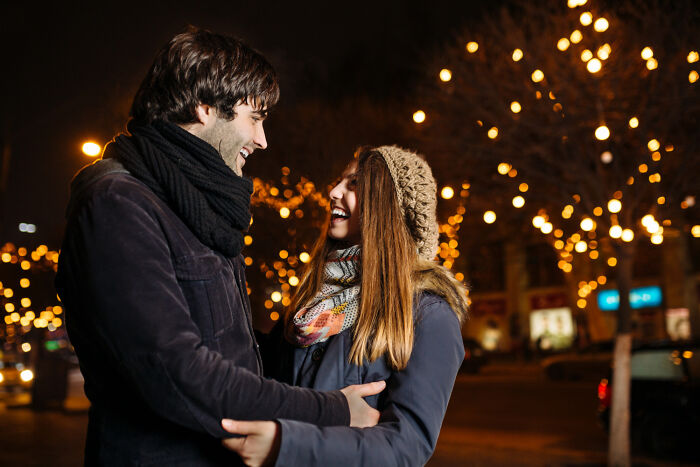 A joyful couple embraces under warm string lights at night, sharing a laugh. This image captures hilarious engagement stories.