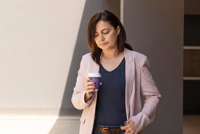 A woman with dark hair in a pink blazer and blue shirt, holding a purple coffee cup, looking down with second-hand embarrassment.