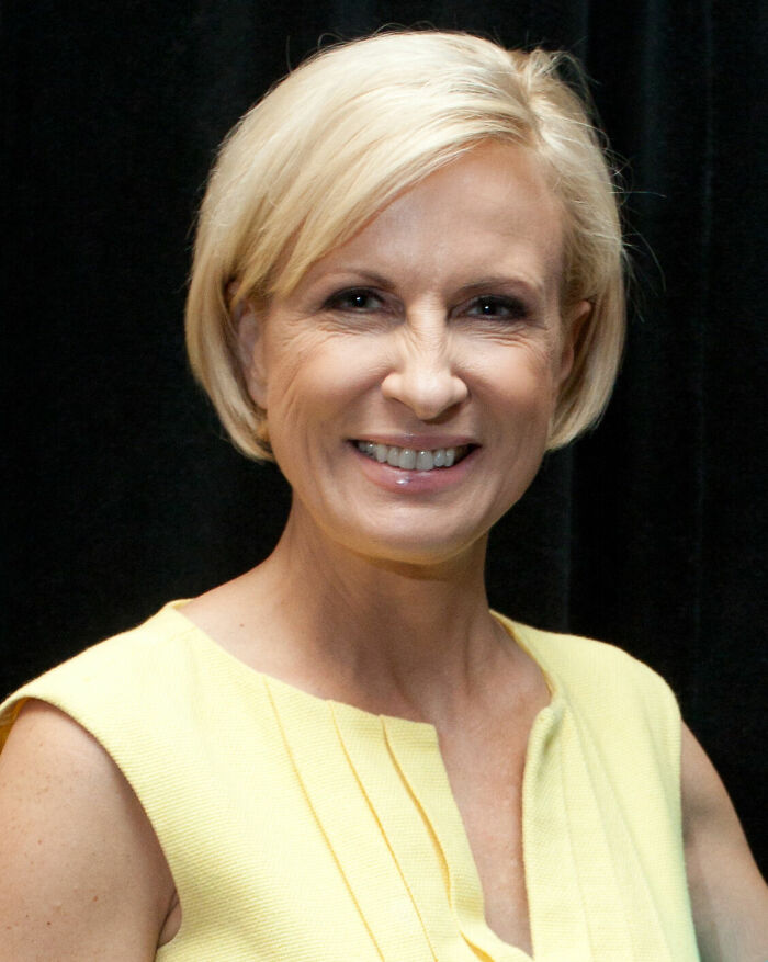 Mika Brzezinski smiling with short blonde hair, wearing a yellow top against a dark background, showcasing her career highlights.