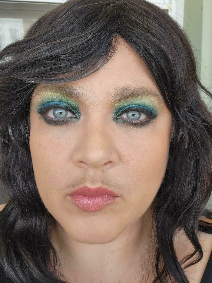 Close-up of a person with dark wavy hair and blue eyes, wearing vibrant green and blue eye makeup. A beauty disaster.