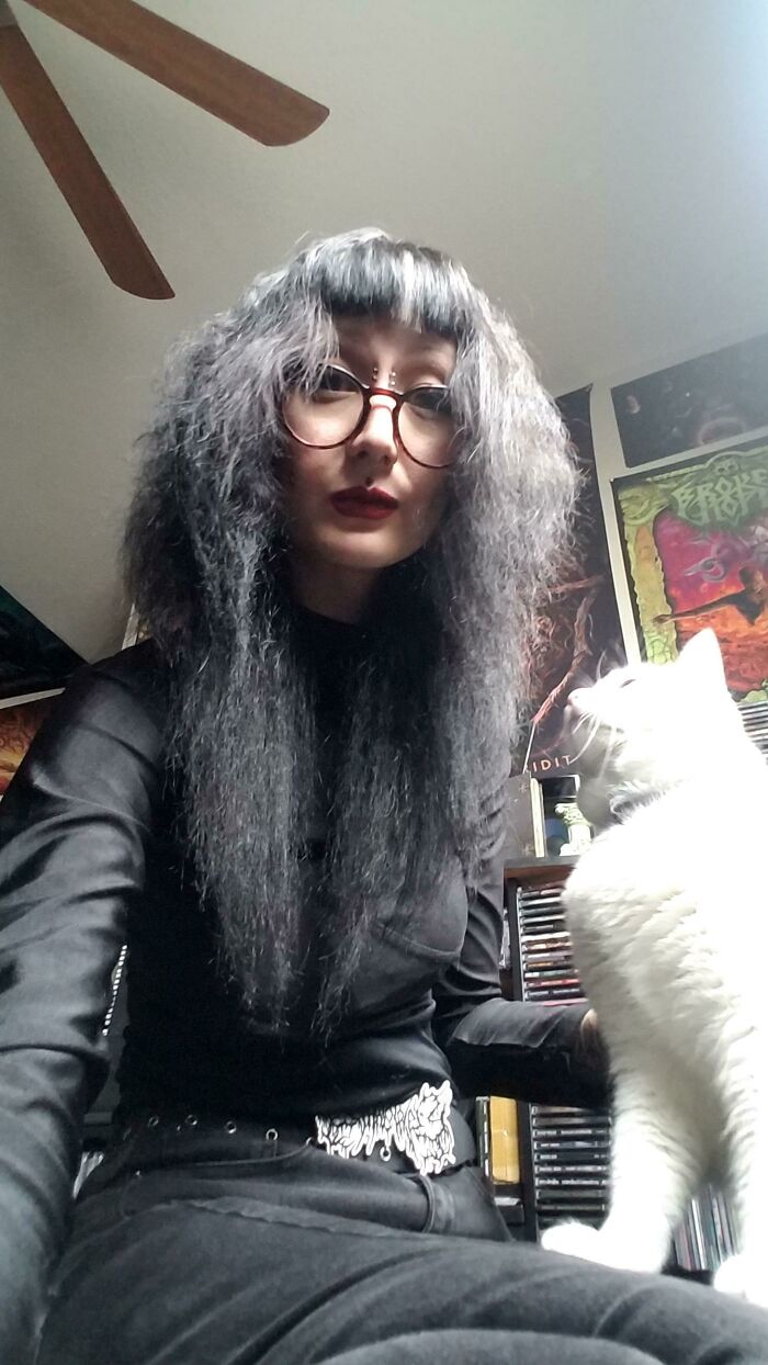 A young woman with curly gray hair, red glasses, and dark lipstick sits beside a white cat. Could be a beauty disaster.