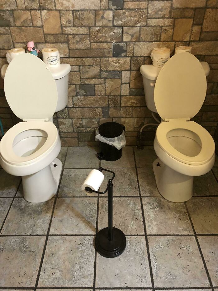 Two toilets side by side with awkwardly placed toilet paper stand