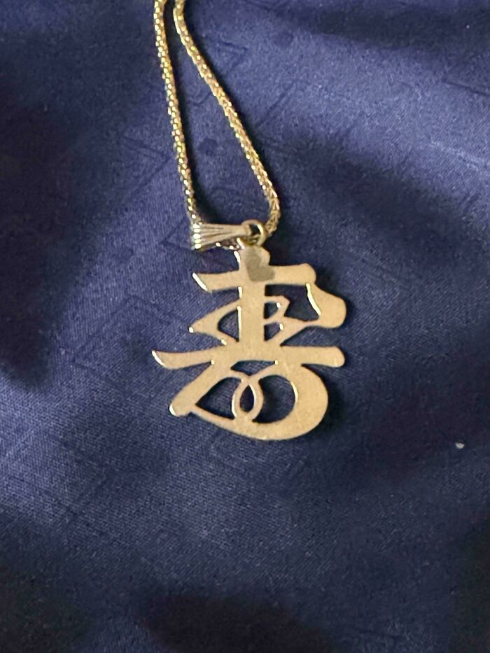 A gold necklace with an interesting Chinese character pendant, lying on a dark blue fabric. What does this mean?