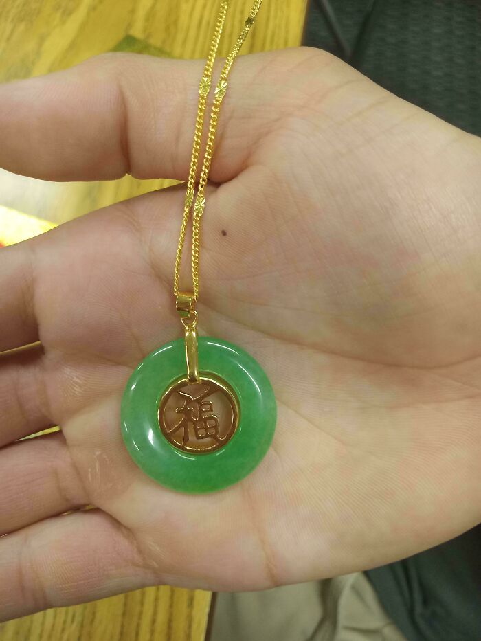 A hand holds a green jade pendant on a gold chain. The pendant has a gold 福 symbol. What does this mean?