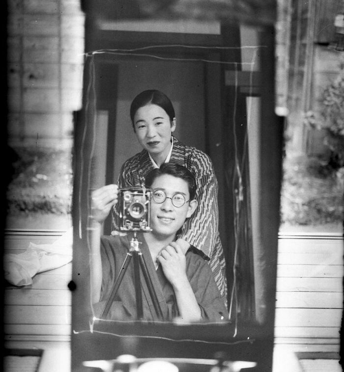 A black and white image of a smiling couple in kimonos, reflected in a mirror. He holds a vintage camera on a tripod. Wholesome pics.