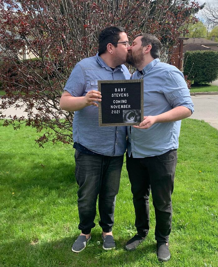 Two men kiss while holding a letter board announcing Baby Stevens arriving Nov 2021, a truly wholesome pic.