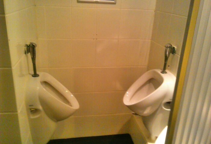 Two urinals placed extremely close in a cramped bathroom corner with poor bathroom design