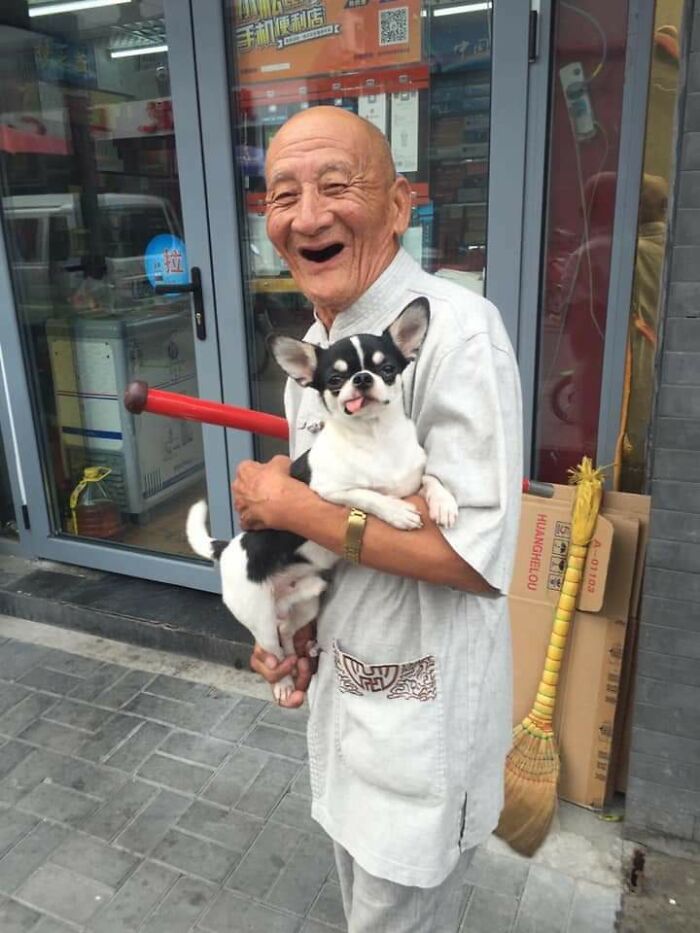 Bald elderly man with a joyful, toothless smile, holding a small Chihuahua with its tongue out. A wholesome pic.