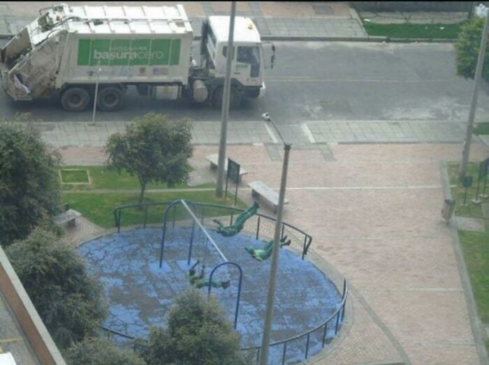 Wholesome pics of a playful art installation: green sculptures swinging upside down in a park, with a garbage truck on the street.