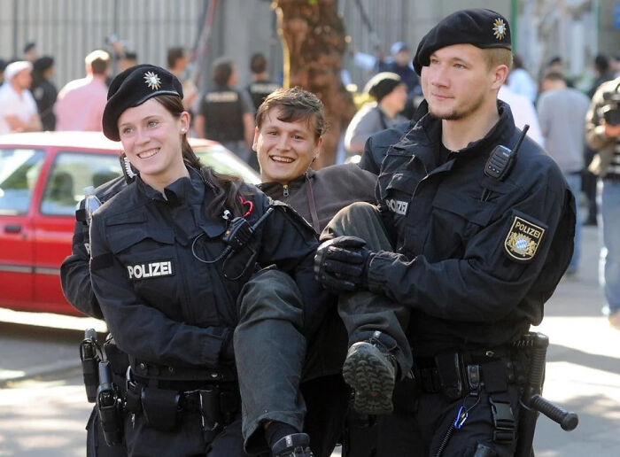 Two smiling police officers in black uniforms and berets carry a laughing man, a wholesome pic.