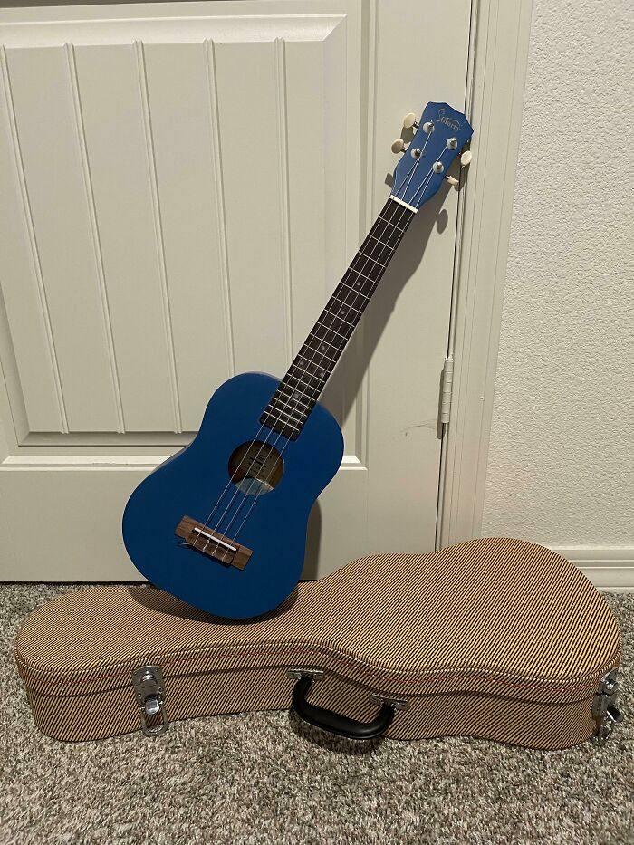 A bright blue ukulele leans against a white door with its tweed case on the carpeted floor. One of many wholesome pics.