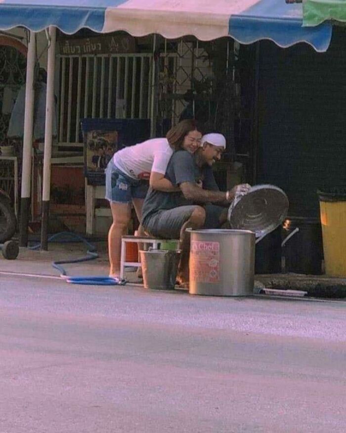 A smiling woman hugs a man washing dishes under an awning; a wholesome pic of everyday support.