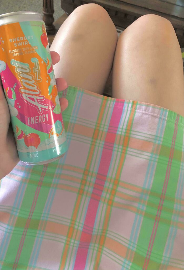 A person holds an Alani Energy Sherbet Swirl can. Its colors accidentally match the pink, orange, and green plaid skirt, creating a satisfying photo.
