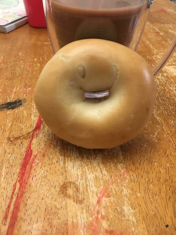 Bagel with a binder clip in the hole, resembling a face, on a wooden table. A wholesome pic of breakfast fun.