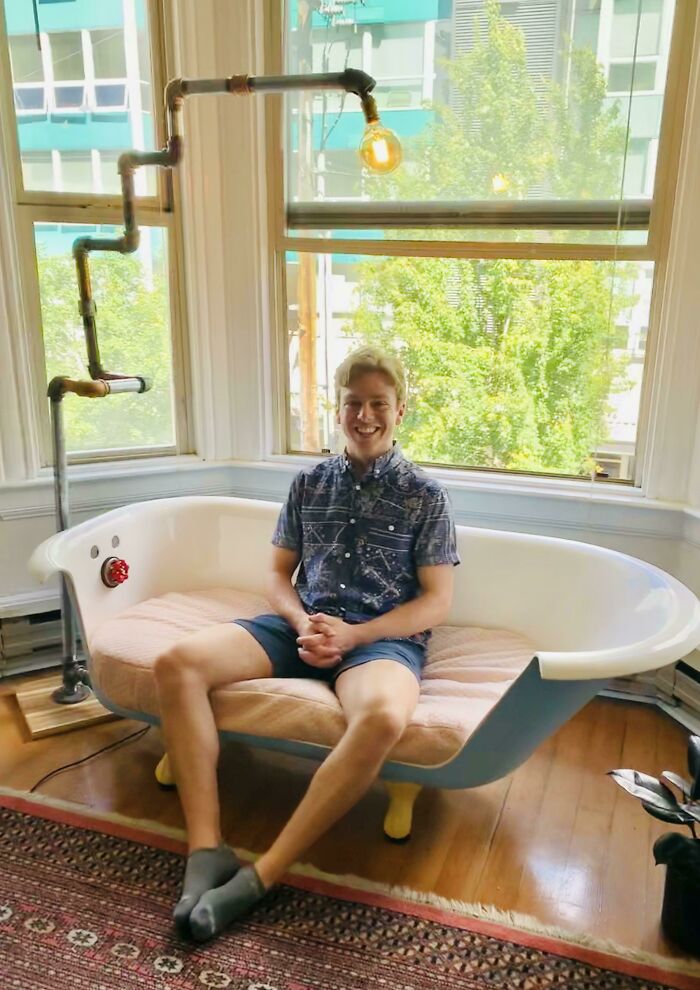 A smiling man sits on a repurposed bathtub couch with a pipe lamp next to a window. A wholesome pic.