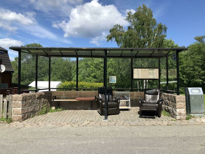 A Wholesome Pic of a cozy bus stop with two armchairs, a bookshelf, and a bench, surrounded by trees under a blue sky.