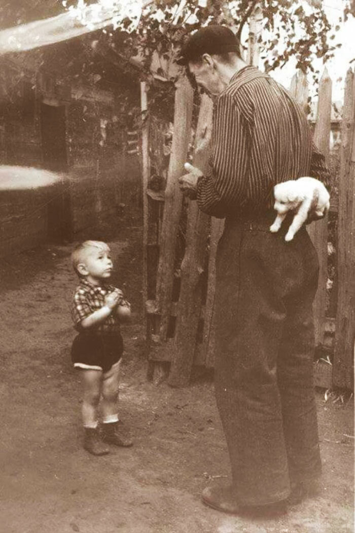 A sepia-toned image of a man holding a tiny white puppy behind his back, surprising a young boy. Wholesome pics.