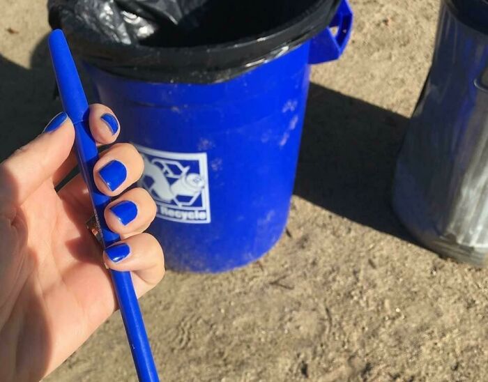A person's hand with blue nail polish holding a blue pen, matching a blue recycle bin, an accidental color match that may lower your cortisol levels.