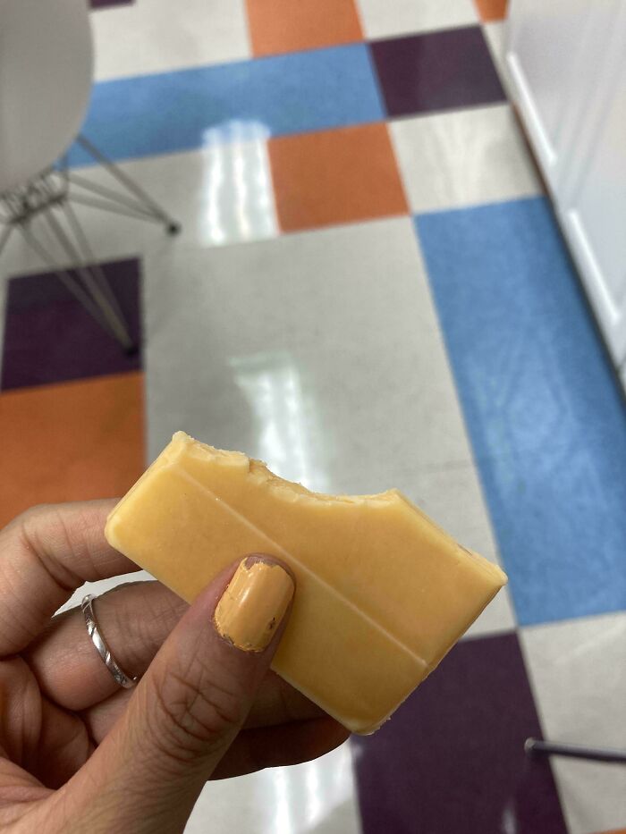 A hand with chipped orange nail polish holds a bitten cheese stick, matching the orange floor tile, a satisfying color match.