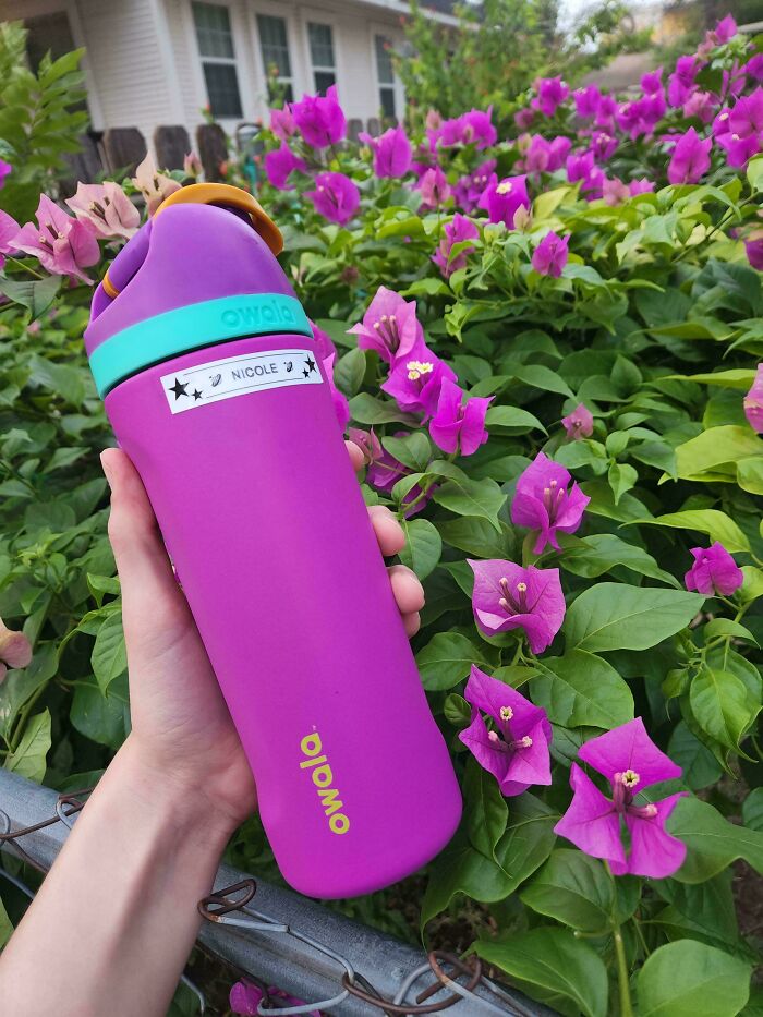 A hand holds a purple Owala bottle with a custom Nicole label, perfectly color-matching vibrant bougainvillea flowers. Accidental color match to lower cortisol levels.