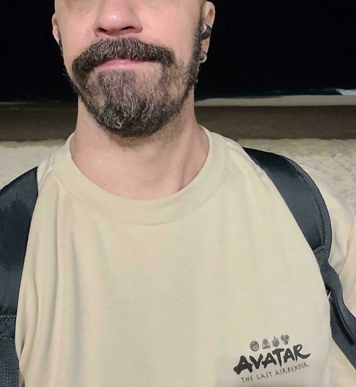 A man with a beard and mustache wearing a light tan Avatar t-shirt, standing on a beach at night. Satisfying color matches.