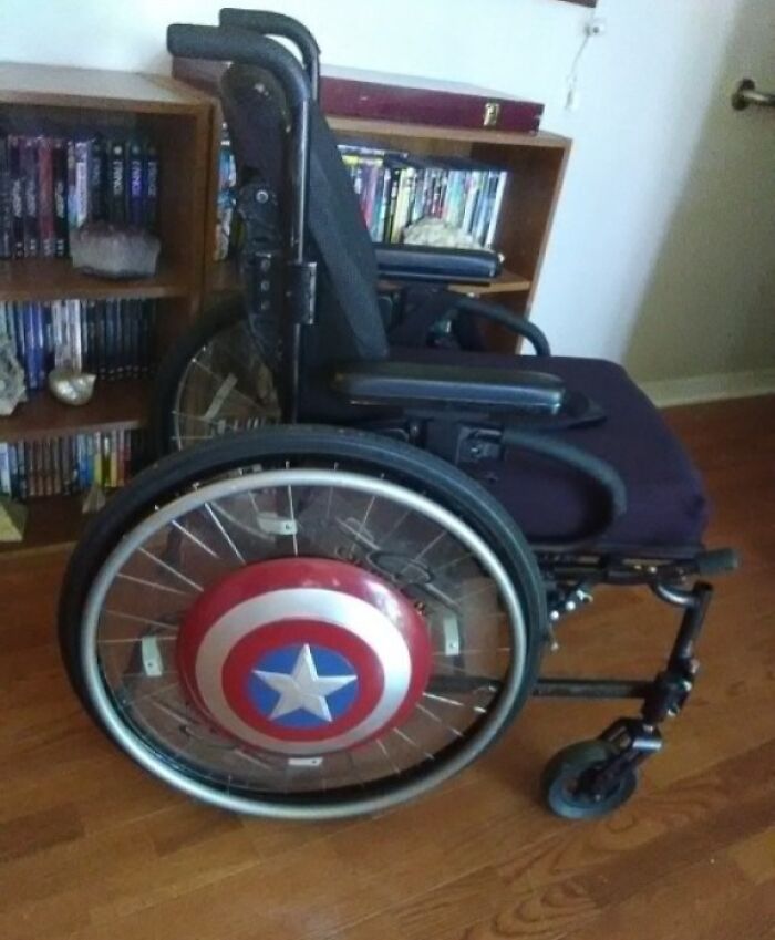 A wheelchair with a Captain America shield on its wheel, next to a bookshelf. Wholesome pics.