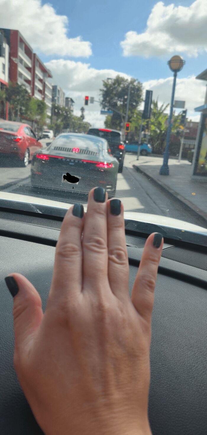 A hand with dark gray nail polish rests on a car dashboard, matching a gray Porsche in traffic. Accidental color matches.