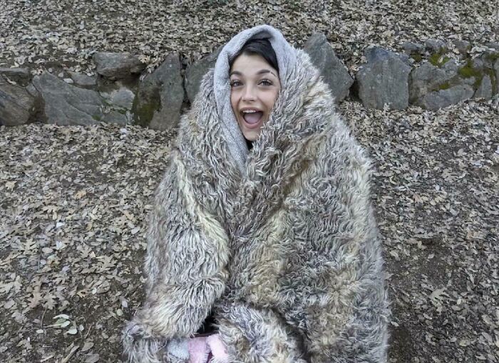 A young woman with an open mouth, wrapped in a fluffy, textured gray blanket, blending with fallen leaves. Satisfying photo.