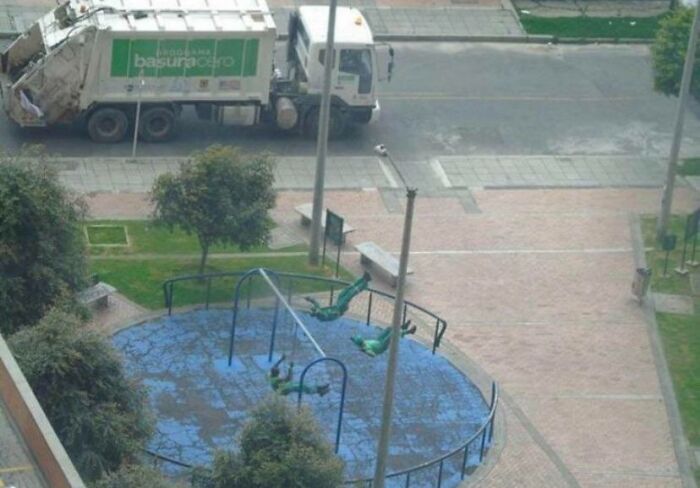 Wholesome pics of a playful art installation: green sculptures swinging upside down in a park, with a garbage truck on the street.