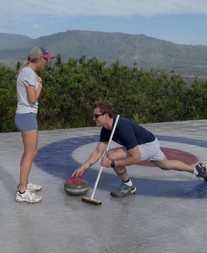 A man makes a curling proposal, kneeling with a curling stone. A woman watches, surprised, outdoors on a sunny day. This proposal pic is hilarious.