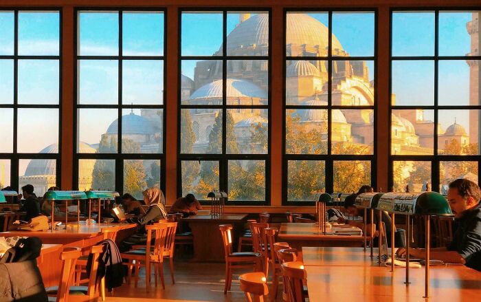 Library interior with large windows overlooking historic building and students studying