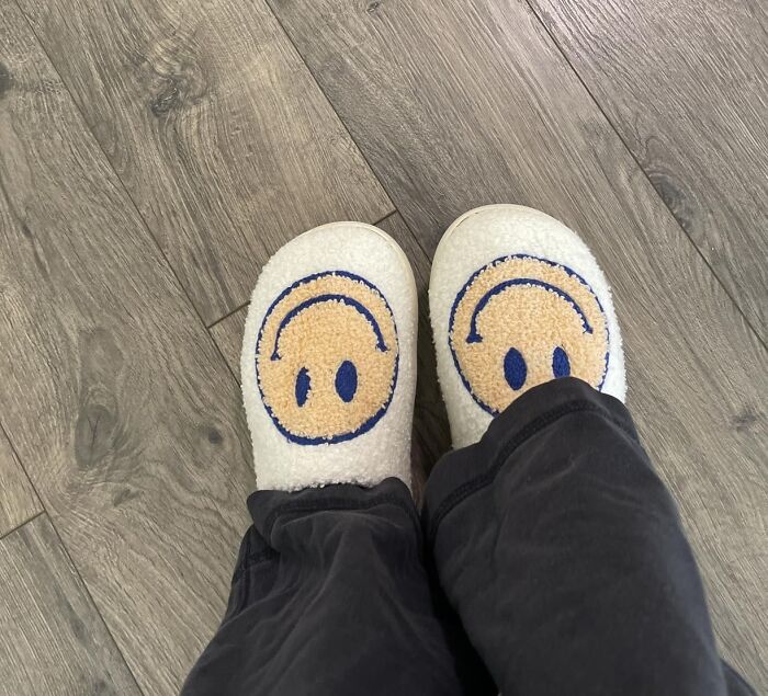 White fuzzy slippers with blue and tan smiley faces, worn on a wood floor. Perfect last minute Mothers Day gifts.
